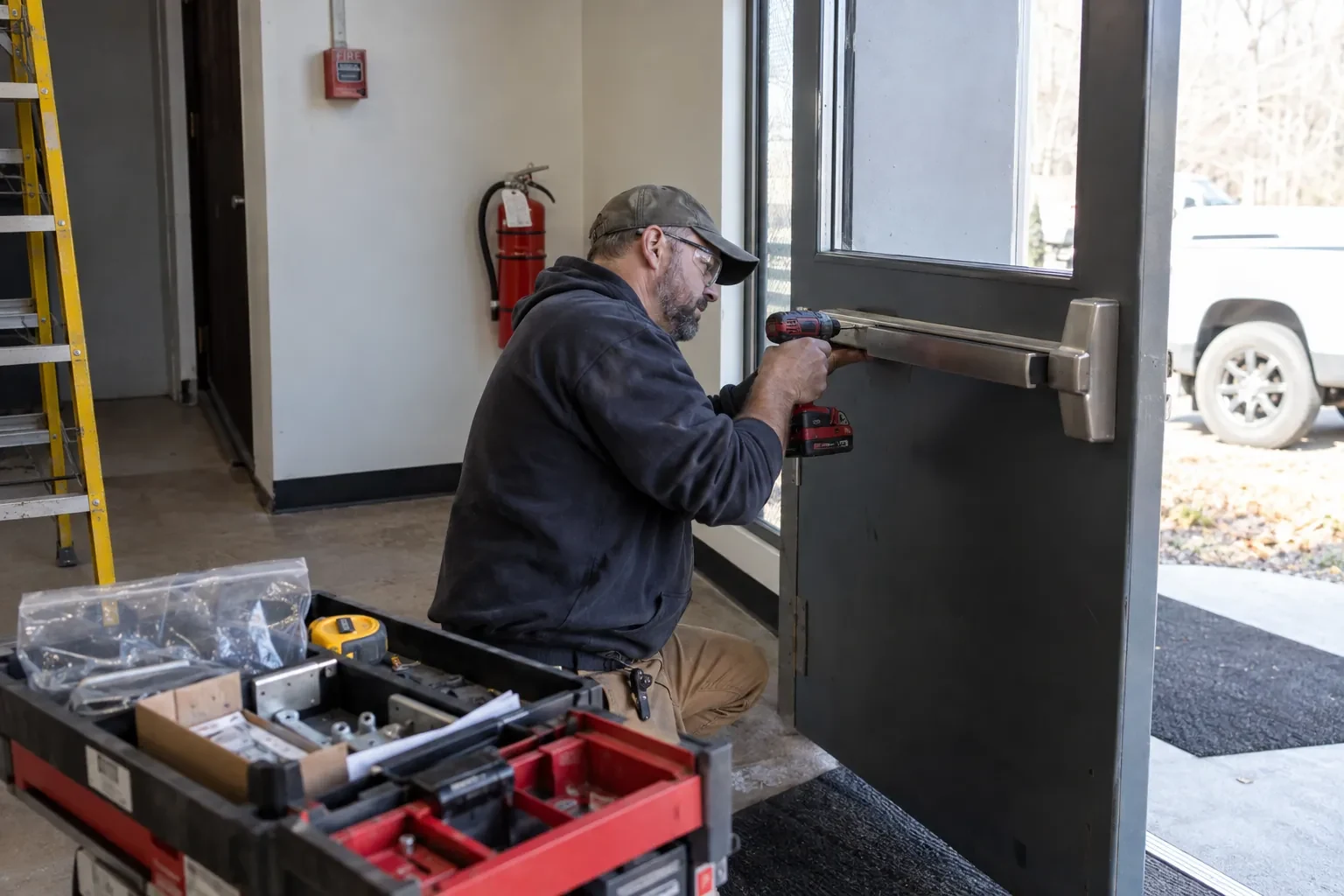 Technician in safety glasses kneeling at a gray commercial door, drilling a steel push bar into place beside an open parts case. — Georgetown