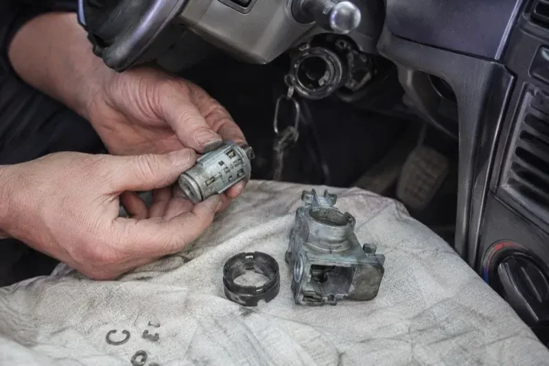 Hands holding a removed ignition cylinder beside a car steering column, with the housing and small parts on a cloth. — Ignition Lock Repair, Halton Hills