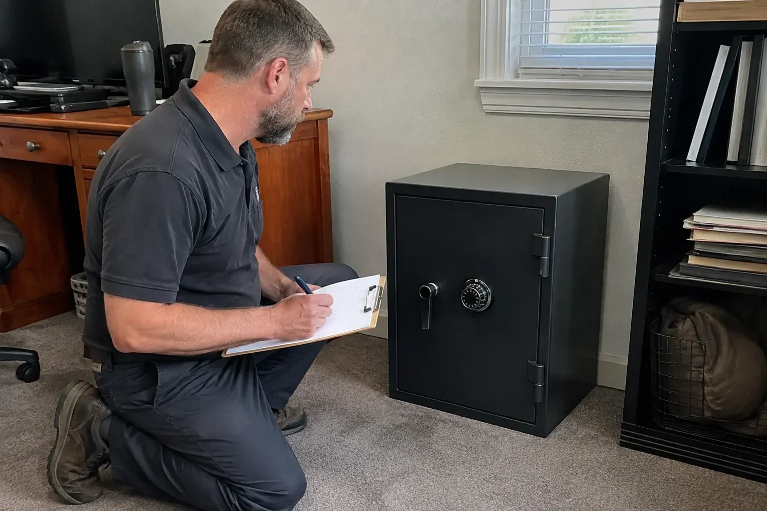 Person writing on a clipboard while kneeling beside a closed black safe with a numbered combination dial in a home office. — Safe Opening & Combination Change, Halton Hills